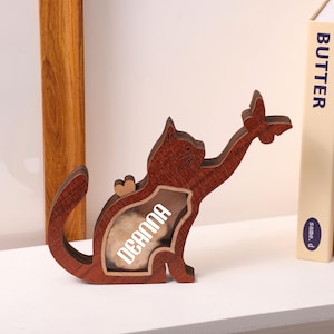 May include: A wooden cat-shaped decorative item with a butterfly and the name "Deanna" visible. The cat is brown with a heart-shaped accent. The item is on a white surface near books.