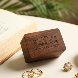 May include: A dark wooden ring box with the engraved names "Powell & Gloria" and the date "17.02.26". The box is next to an open book, gold rings, and pearl earrings. The box is rectangular with a hinged lid.