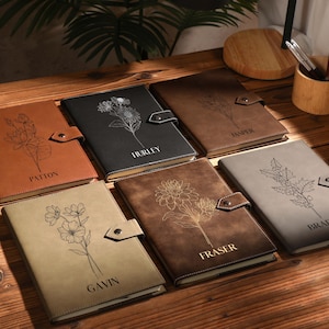 May include: A selection of personalised journals in shades of brown, black, and grey. Each journal displays a floral design and a name engraved on the cover. The journals are secured with a button clasp.