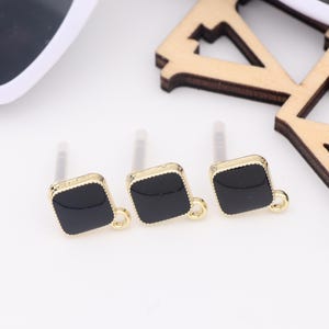 May include: Three gold-toned square charms with black enamel centers and a small loop on each side. The charms are on a white surface.
