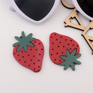 May include: Two red and green strawberry shaped resin embellishments with white polka dots.