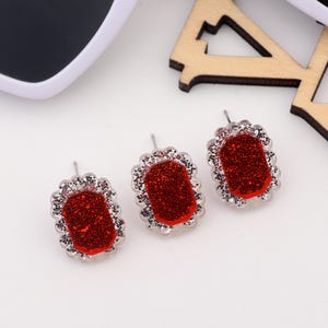 May include: A pair of red glitter earrings with silver accents. The earrings are shaped like a rectangle with rounded corners.