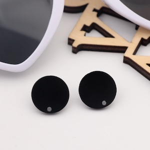 以下が含まれることがあります： 中央に小さな穴が開いた黒い丸いイヤリングが2つ。イヤリングは白い表面の上に置かれています