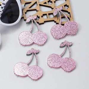 May include: Four pink glitter cherry charms with green stems and leaves. The charms are arranged on a white surface with a pair of sunglasses and a wooden decorative piece in the background. These charms are ideal for jewelry making or crafting.