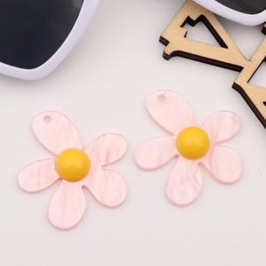 May include: Two pink resin daisy flower charms with yellow centers. The charms have a hole at the top for attaching to jewelry or other crafts.