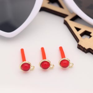 Puede incluir: Tres hallazgos de joyería de color rojo y dorado. Cada hallazgo tiene una cuenta redonda roja engastada en un bisel dorado, con un pequeño lazo y un tallo rojo. Gafas de sol y un recorte de madera en el fondo.