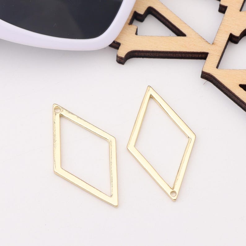 Op de afbeelding: Twee gouden metalen hangers in de vorm van een diamant met een klein gaatje bovenaan om aan sieraden te bevestigen.