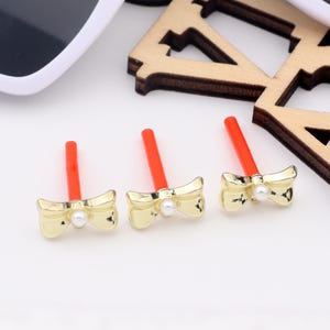 May include: Three gold bow-shaped charms with a pearl in the centre, each attached to a red stick.