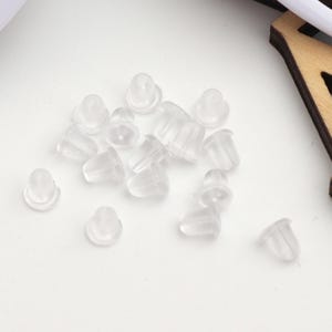 Puede incluir: Primer plano de un montón de respaldos de pendientes de plástico transparente. Los respaldos son pequeños y redondos, con un pequeño agujero en el centro. Están diseñados para ser usados con pendientes de botón.