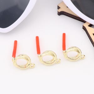May include: Three gold-toned metal ring clasps with red plastic inserts. The clasps are designed to be used for jewelry making.