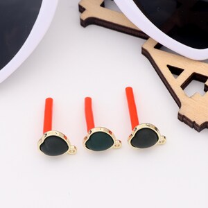 May include: Three gold-colored heart-shaped charms with green centers and orange stems. The charms are arranged in a row on a white surface.