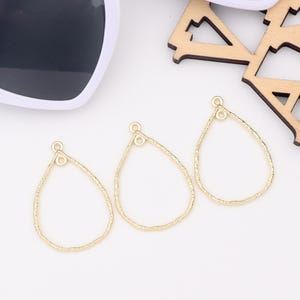 Könnte beinhalten: Drei goldfarbene, tropfenförmige Ohrringanhänger mit strukturierten Kanten. Jeder Anhänger hat oben eine kleine Schlaufe zum Befestigen an Ohrringhaken. Die Anhänger werden auf einer weißen Oberfläche präsentiert.
