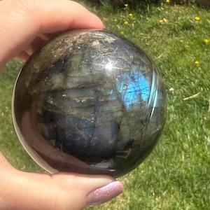 May include: A polished labradorite sphere, held in a hand, displays a captivating play of color. The sphere's surface showcases iridescent flashes of blue, green, and gold, set against a backdrop of dark gray and black. The sphere is a decorative item.