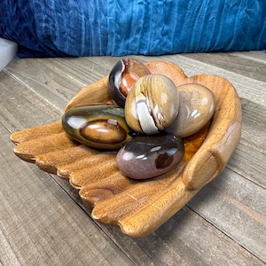May include: A wooden hand-shaped bowl holds five polished stones. The stones are various colors, including brown, tan, and black. The bowl is made of light brown wood and sits on a wooden surface.