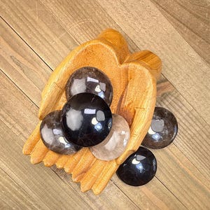 May include: A carved wooden bowl shaped like two cupped hands holding several smooth, polished, round stones in shades of translucent black, dark gray, and clear quartz. The stones are varied in size and have a glossy, reflective surface.