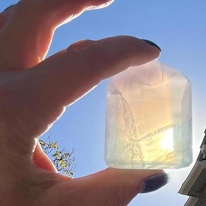 May include: A clear, iridescent crystal with a light pink hue. The crystal is shaped like a rectangular prism and is held in a hand against a bright blue sky.