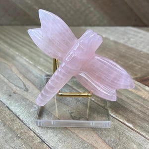 May include: A pink rose quartz dragonfly sculpture on a gold stand. The dragonfly is carved with intricate details and has a delicate, feminine feel.