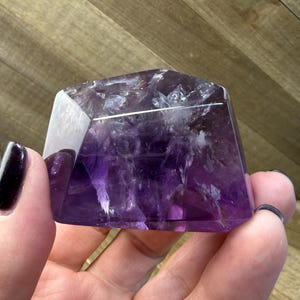 May include: A faceted amethyst crystal with a polished surface. The crystal is a deep purple color with white and clear inclusions. The crystal is held in a hand.