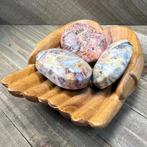 May include: A wooden hand-shaped bowl holds three polished stones. The stones have a variety of colors, including red, gray, and yellow. The bowl is made of light brown wood and sits on a wooden surface.