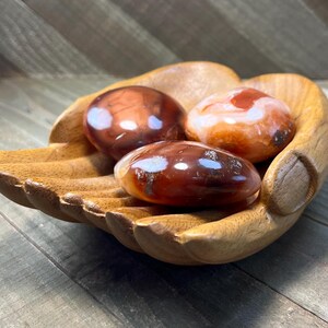 May include: Three carnelian tumbled stones in a wooden hand-shaped bowl. The stones are a mix of red and orange with white banding.
