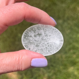 May include: A clear, oval-shaped crystal with a textured surface. The crystal is held in a hand with purple nail polish.