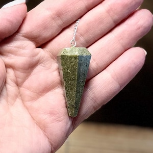 May include: A faceted, olive-green pendulum with a silver chain. The gemstone is held in a hand, showcasing its geometric shape and textured surface. The pendulum is designed for divination and spiritual practices.