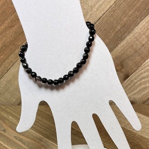 May include: Black gemstone bracelet with a single clear gemstone accent. The bracelet is displayed on a white hand-shaped stand.