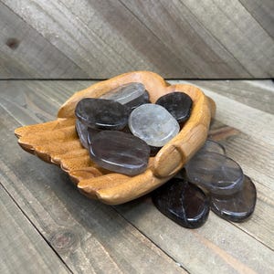 May include: A carved wooden bowl shaped like a pair of cupped hands holding a collection of smooth, polished, oval-shaped stones. The stones vary in color from translucent clear to dark smoky gray and black, with some resting inside the bowl.