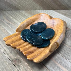 May include: A wooden hand-shaped bowl holds several polished, oval-shaped stones. The stones are a deep teal color with darker green inclusions. The bowl is made of light-colored wood with visible grain.
