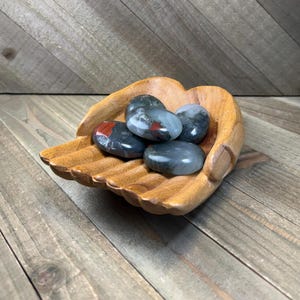 May include: A carved wooden bowl shaped like two cupped hands holding a collection of smooth, polished stones. The stones are primarily dark gray and black with distinct patches of deep red and white mineral inclusions throughout the surfaces.