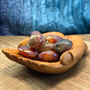 May include: A wooden hand-shaped bowl filled with iridescent agate tumbled stones. The stones are a mix of pink, orange, and brown with a shimmering, iridescent finish.