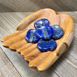 May include: A wooden hand-shaped bowl holds five polished, heart-shaped lapis lazuli stones. The stones are a deep blue with streaks of lighter blue and gray. The bowl is made of light brown wood.