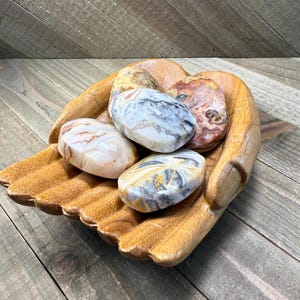 May include: A wooden hand-shaped bowl filled with five polished stones. The stones are oval-shaped and feature a variety of colors, including white, gray, yellow, and red. The bowl is made of light brown wood.