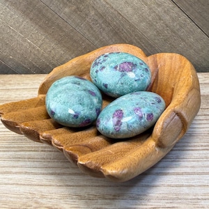 May include: A carved wooden bowl shaped like two cupped hands holding three smooth, polished stones. The stones are a mottled teal green color with distinct, irregular patches of deep magenta and dark purple throughout their surfaces.