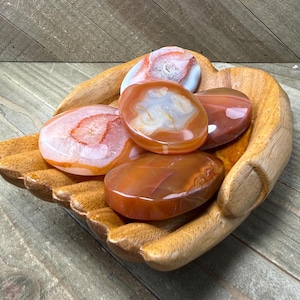 May include: A wooden hand-shaped bowl filled with polished agate stones. The stones are various shades of orange, red, and white. The bowl is made of light brown wood and sits on a wooden surface.