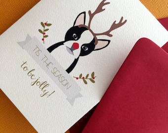 May include: A white card featuring a black and white dog with a red nose and antlers. The card reads "Tis the season to be jolly!"