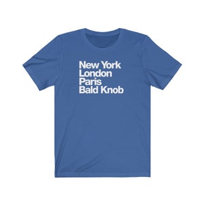 May include: A royal blue t-shirt with white text that reads "New York London Paris Bald Knob".
