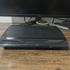 May include: A black Sony PlayStation 3 game console featuring a ribbed top surface and a smooth front panel with the Sony logo on the left side and the PlayStation logo on the right. The console has a rectangular shape with visible surface scratches.