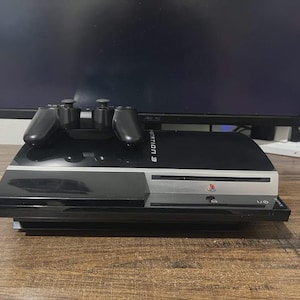 May include: A black PlayStation 3 game console with a glossy finish and a silver front panel featuring the PlayStation logo. A black wireless game controller rests on top of the console, which is positioned in front of a black computer monitor.