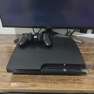 Puede incluir: Una consola de videojuegos Sony PlayStation 3 negra con un mando inalámbrico negro a juego colocado encima. La consola presenta el logotipo de Sony en la parte frontal izquierda y el logotipo de PS3 en relieve en la parte superior, frente a un monitor.