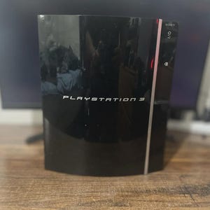 Pode incluir: Uma consola de jogos Sony PlayStation 3 preta com acabamento brilhante. O painel frontal apresenta o texto PLAYSTATION 3 em letras brancas, juntamente com um friso vertical prateado, um botão de energia e um botão de ejeção no lado direito.