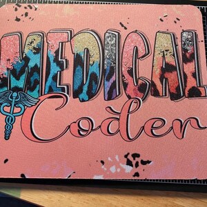 Könnte beinhalten: Ein rechteckiges Mauspad mit pfirsichfarbenem Hintergrund und schwarzen Farbspritzern. Der Text "MEDICAL Coder" ist in einem bunten Leopardenmuster gedruckt, begleitet von einem blauen Caduceus-Symbol auf der linken Seite.
