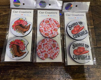 May include: Three sets of two circular car coasters in clear plastic packaging. The first set features brown cowboy boots with red hats and flowers. The second set has a pink pattern of boots, horseshoes, and hearts. The third set shows red hats with the text "Saddle Up Cowgirls."
