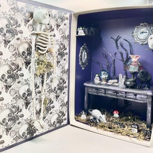 May include: A miniature diorama with a skeleton, a table with a crystal ball, and a rat in a spooky forest setting. The diorama is inside a box with a purple interior and a skull patterned exterior.