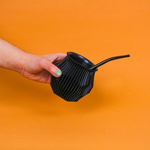 May include: A black mate gourd with a black bombilla straw. The gourd has a ribbed texture and is held in a hand.