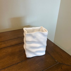 Puede incluir: Jarrón de cerámica blanco con un diseño texturizado y arrugado. El jarrón tiene forma cuadrada y un acabado liso y mate.