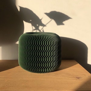 May include: A dark green, cylindrical vase with a wavy, textured surface. The vase sits on a wooden surface, with shadows of leaves cast on the wall behind it. The vase is approximately 8 inches in diameter and 7 inches tall.