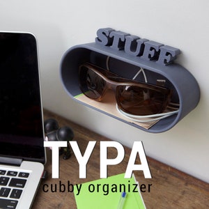 May include: A gray wall-mounted organizer with the word "STUFF" printed on it. The organizer has a small shelf for storing items like sunglasses and cords. The organizer is labeled "TYPA cubby organizer".