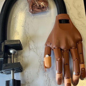 May include: A brown articulated practice hand for nail art, with attached flexible arm and clamp. The hand has tan-colored nail tips and is positioned against a white marble-patterned surface. A bag of additional nail tips is visible.