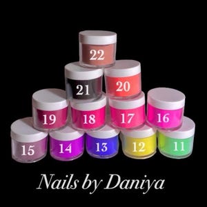 Può includere: Una collezione di piccoli vasetti trasparenti con coperchi bianchi, ciascuno riempito con un colore diverso di polvere per unghie. I vasetti sono numerati da 11 a 22. Il testo "Nails by Daniya" è in basso.